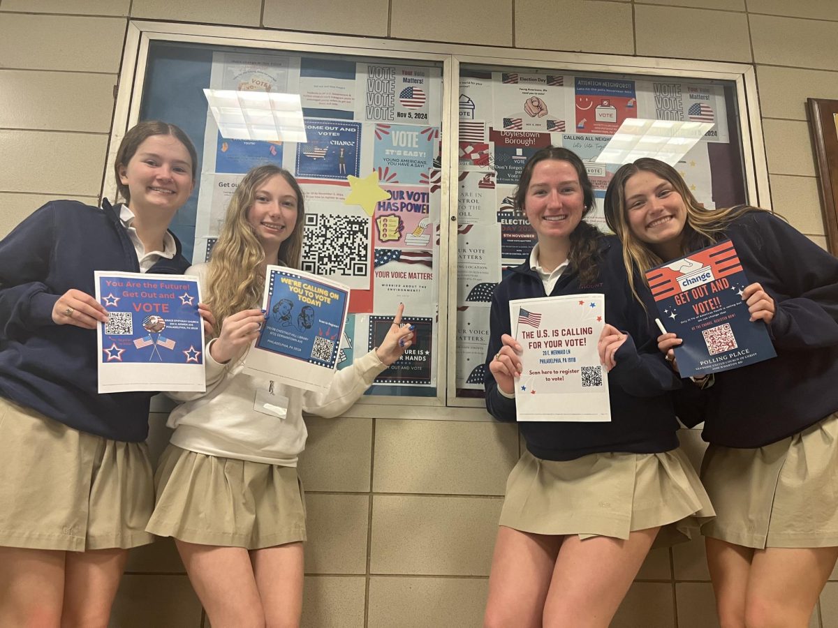 Abby Williams 25, Summer Delli Carpini 25, Grace Remus 25, and Sabrina Carpenter 25 pose in front of a display while holding up flyers they made for Mrs. Feilkes Honors Government class. The project required that students include a QR code on where to register or where to vote. 